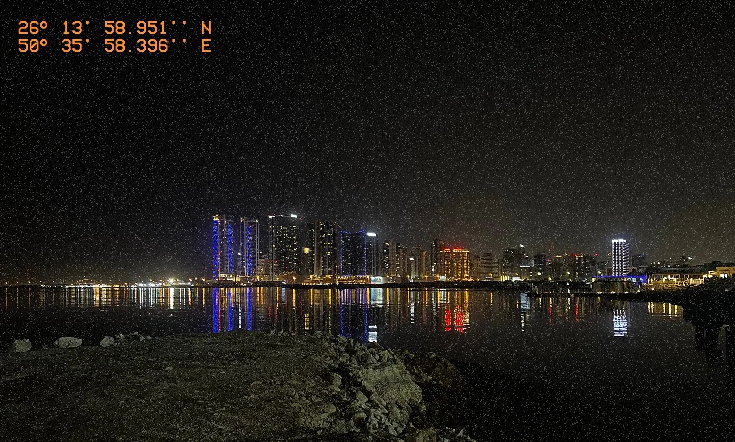 Nighttime cityscape of Juffair with high-rise buildings reflecting on the water, viewed from a rocky shoreline. The GPS coordinates 26°13′58.951″N, 50°35′58.396″E are overlaid in orange text.