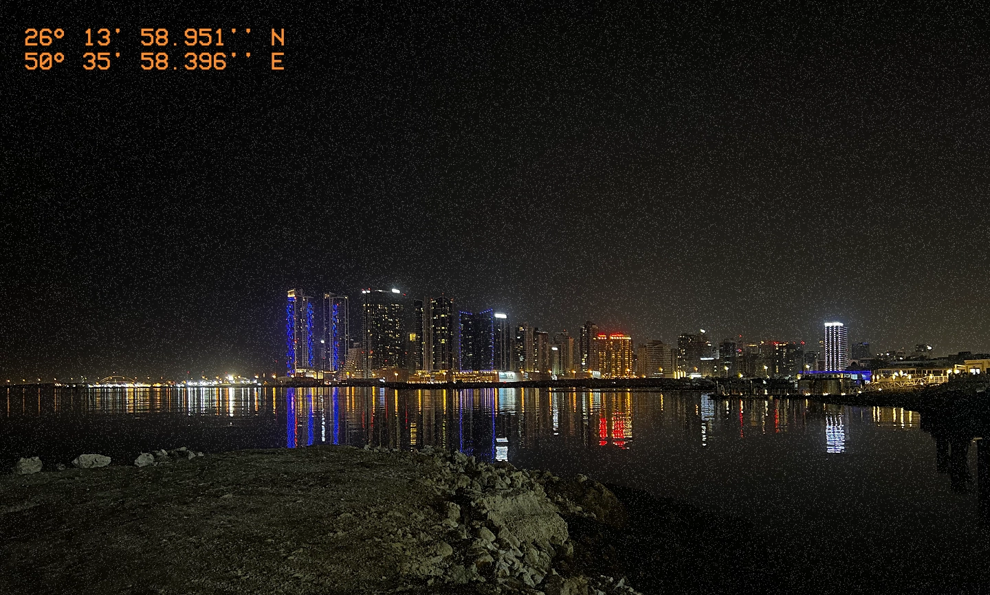 Nighttime cityscape of Juffair with high-rise buildings reflecting on the water, viewed from a rocky shoreline. The GPS coordinates 26°13′58.951″N, 50°35′58.396″E are overlaid in orange text.