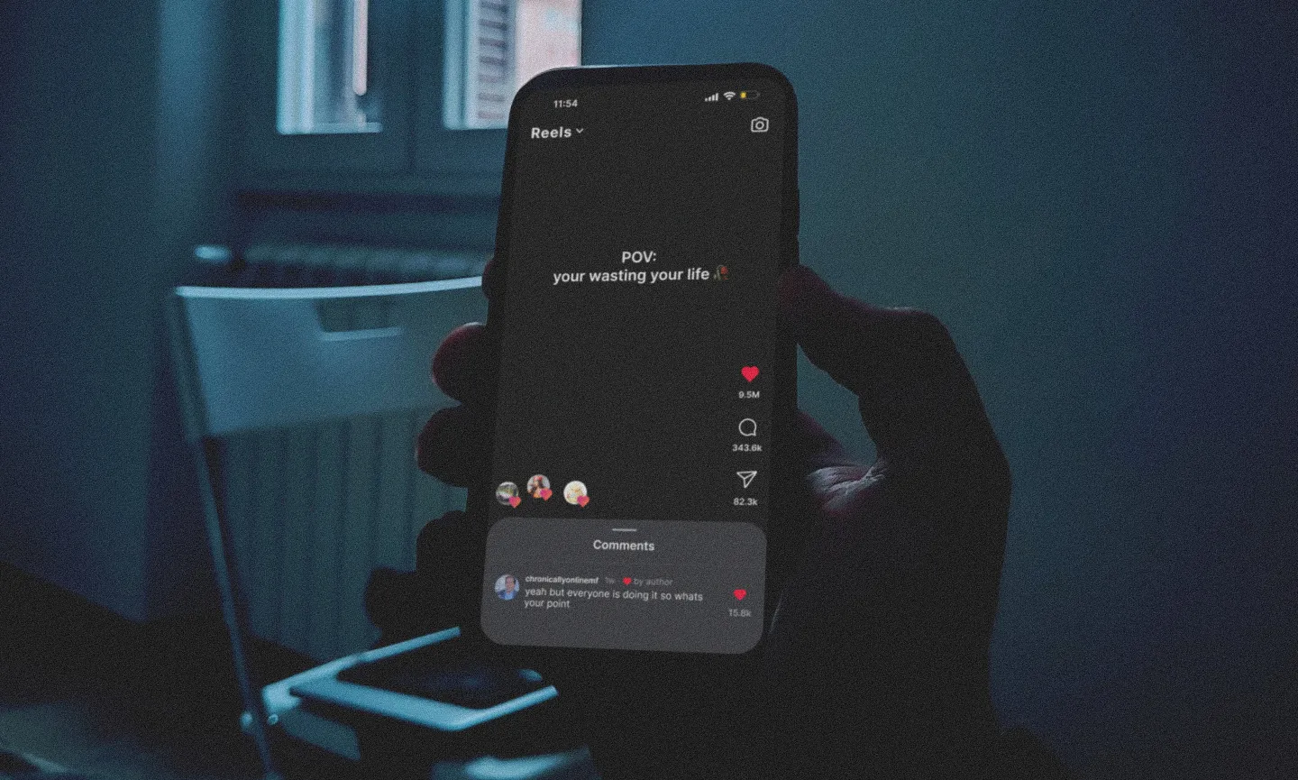 A person holds a smartphone in a dark room, displaying a social media post that reads "POV: your wasting your life".