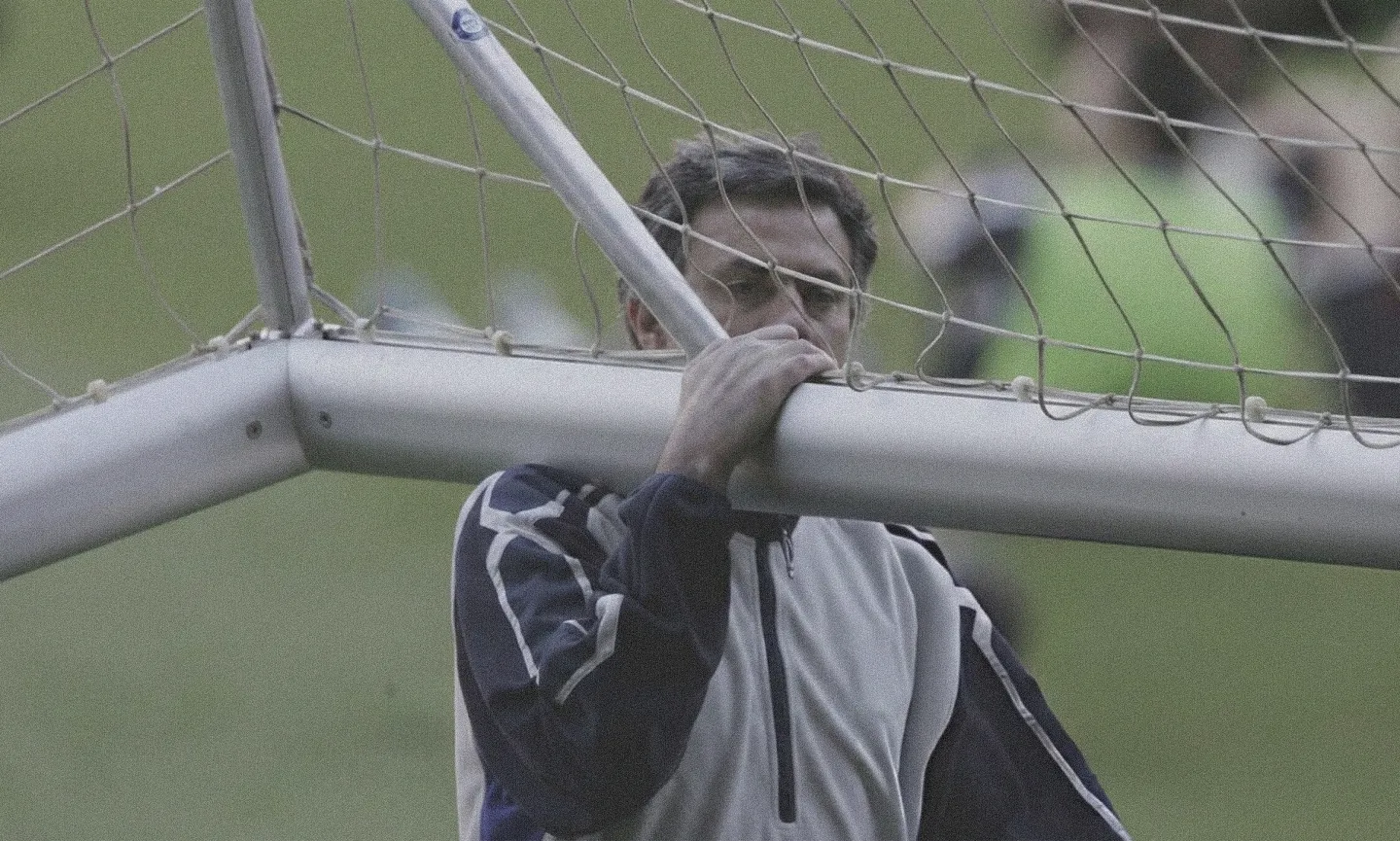 Football manager in a navy and grey tracksuit, looking pensive as he holds onto the crossbar of a football goal.