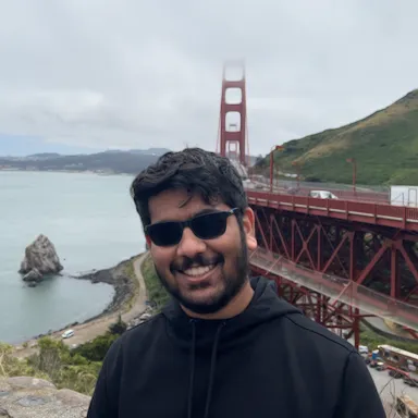 Kelvin DeCosta wearing sunglasses and a black hoodie, smiling in front of the Golden Gate Bridge on a cloudy day.